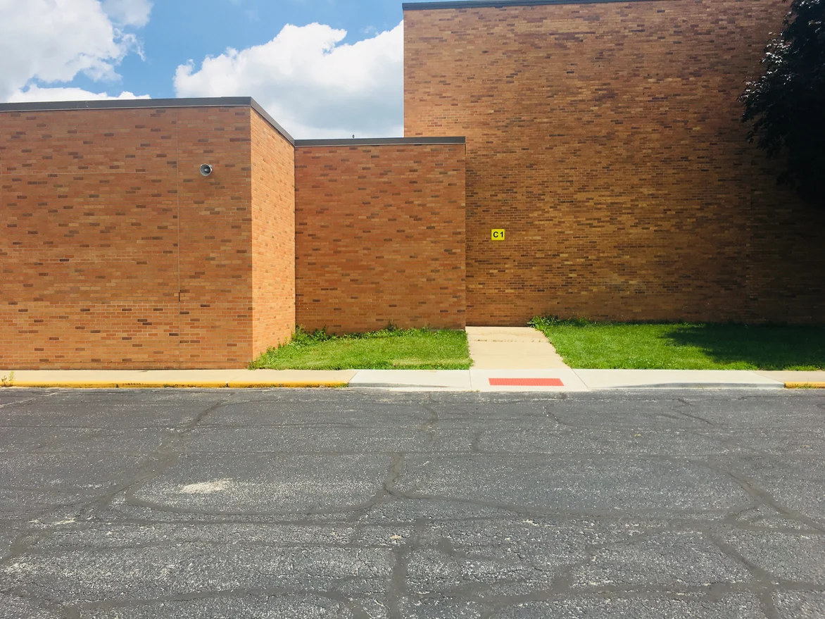 Two blank brick walls of the school meeting at a step in the plan in 2019, a walkway running between them past a door number plate
