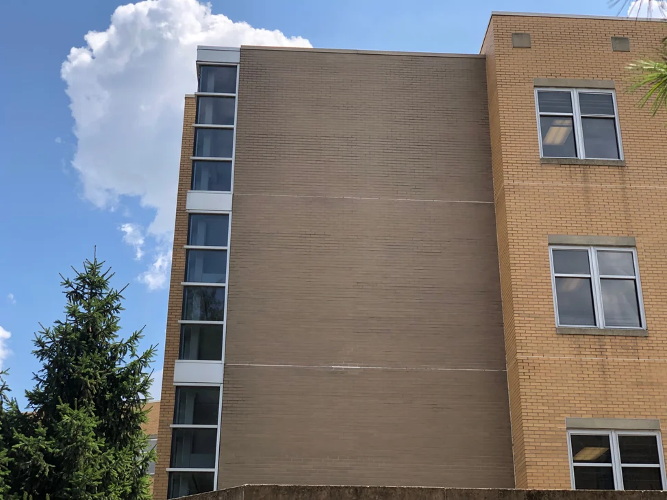 The hospital wall in 2019, one tan range running the full height beside the glazed stair tower, with the buff brick of the wing stepping forward at the right.