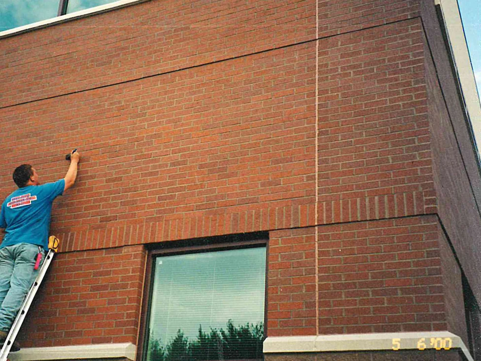 An applicator on a ladder brushing stain onto single bricks in a wall panel, lighter orange units scattered through the red around them