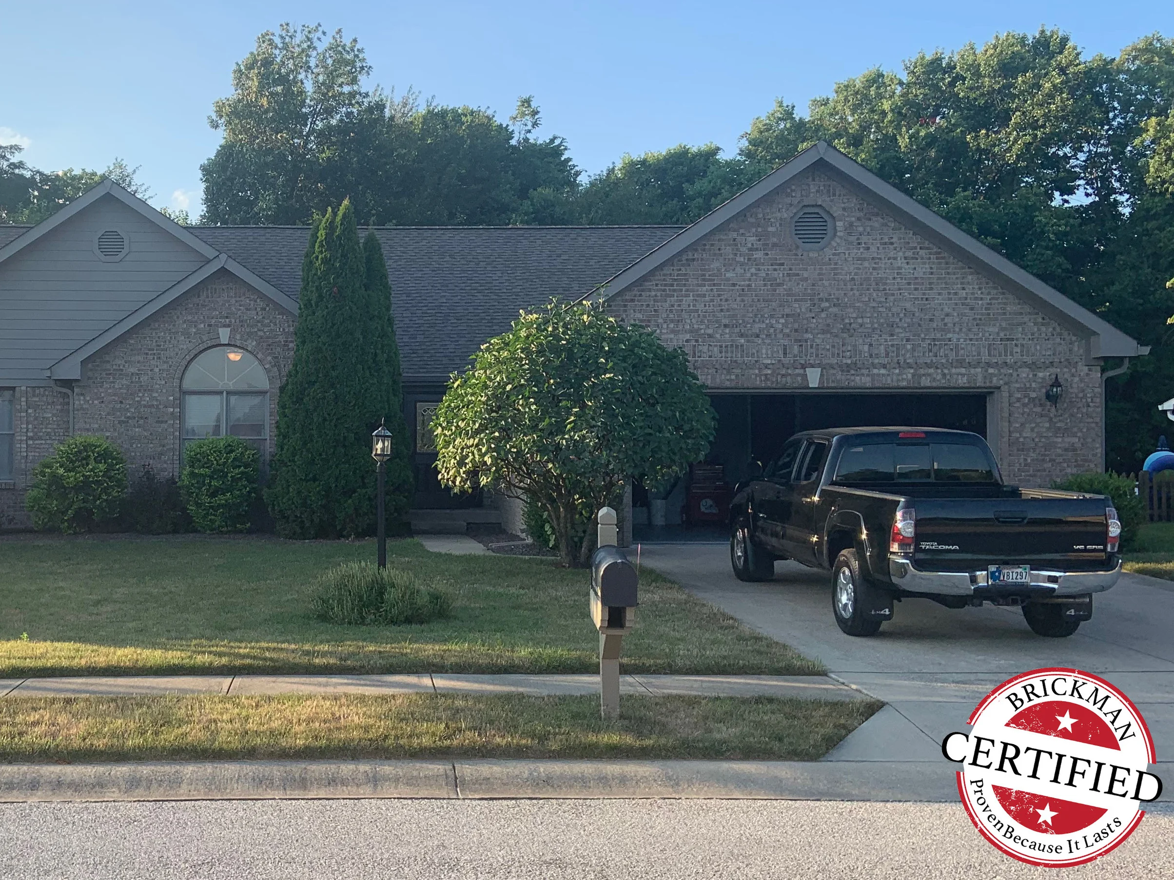 The front of the house from the street in 2019, the entry gable, the arched window and the garage wing all carrying the same even light tan brick, with the certification stamp at the corner of the frame
