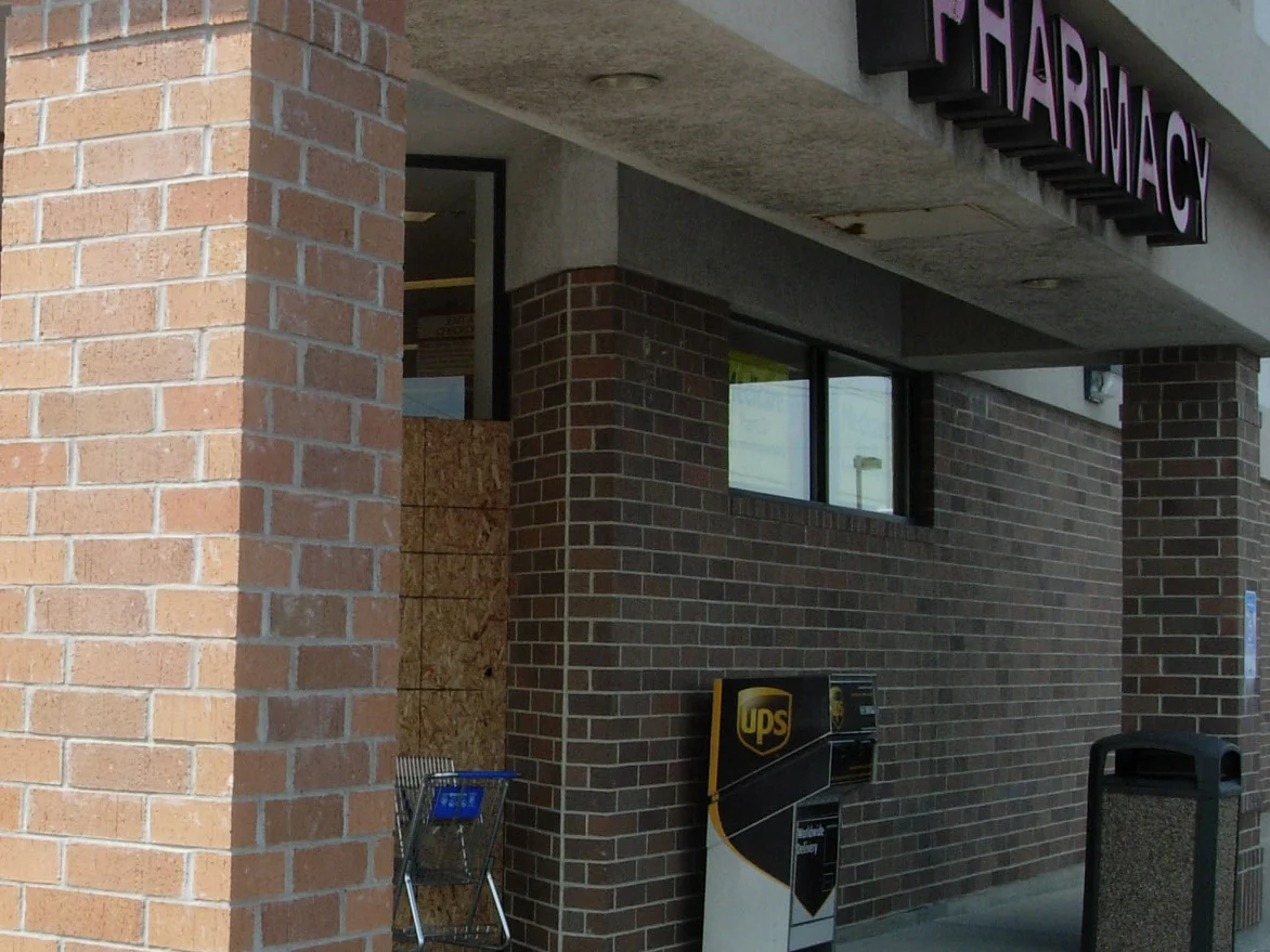 A brick column at a store entrance in pale tan, standing against the darker red brown brick of the wall behind it, with a plywood panel over a damaged section