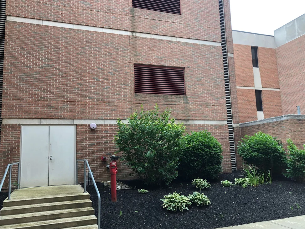 A tall university wall in 2019 with two louvred vents and cast stone banding, its brick reading red and brown in the same range as the wing standing behind it.