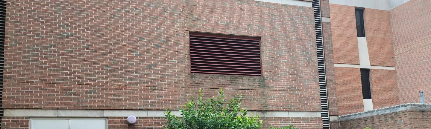 The university building's brick wall in 2019, red and brown across a louvred vent and a course of cast stone banding