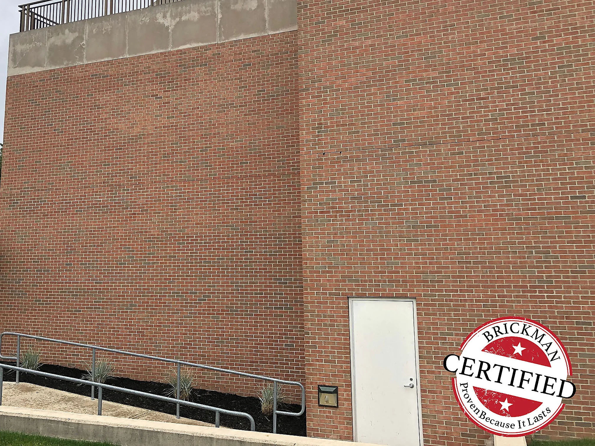 A blank end wall below a rooftop terrace railing in 2019, its red brown brick running evenly from the parapet down to the ramp and the service door at the base