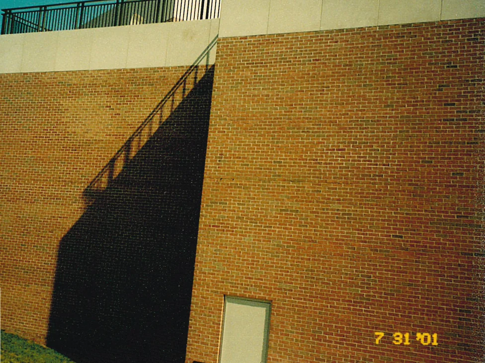 A blank end wall below a rooftop terrace railing in 2001, a long diagonal shadow falling across its left half and a service door at the base of the taller section