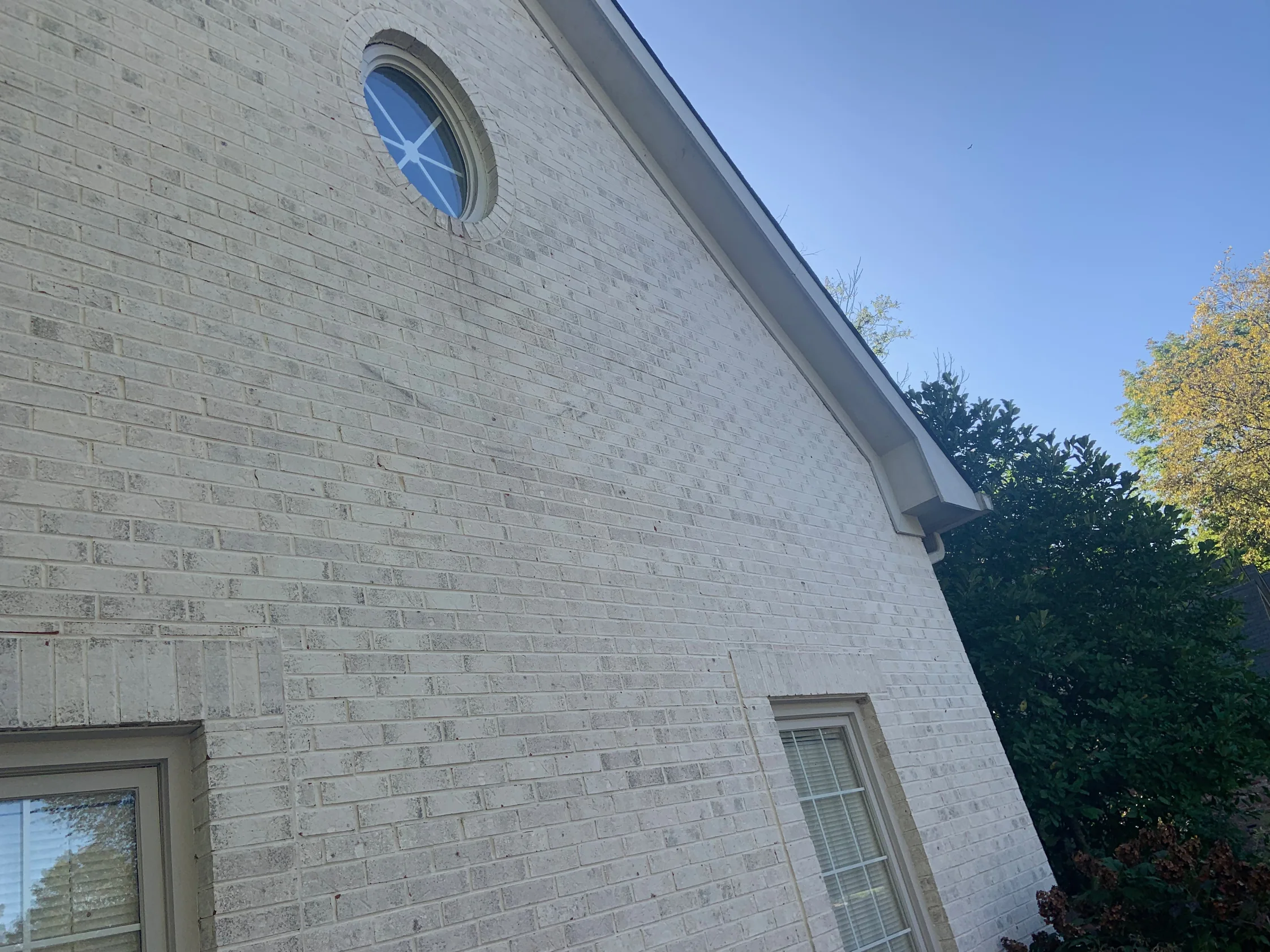 The gable end of the house from below in 2019, the oval window set in brick that reads as one cream and grey range up to the eaves