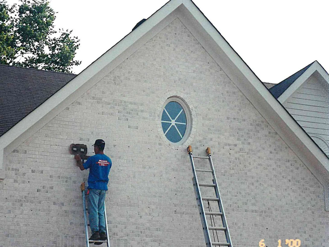 An applicator on a ladder working stain into the gable end of a cream brick house, an oval window set at the centre of the gable