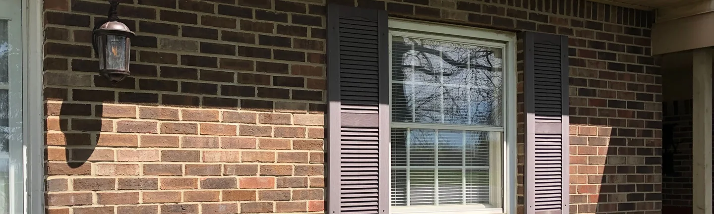 A brick wall beside a front door in 2019, a lamp above and a shuttered window to the right, the lower courses running lighter than the dark brick above
