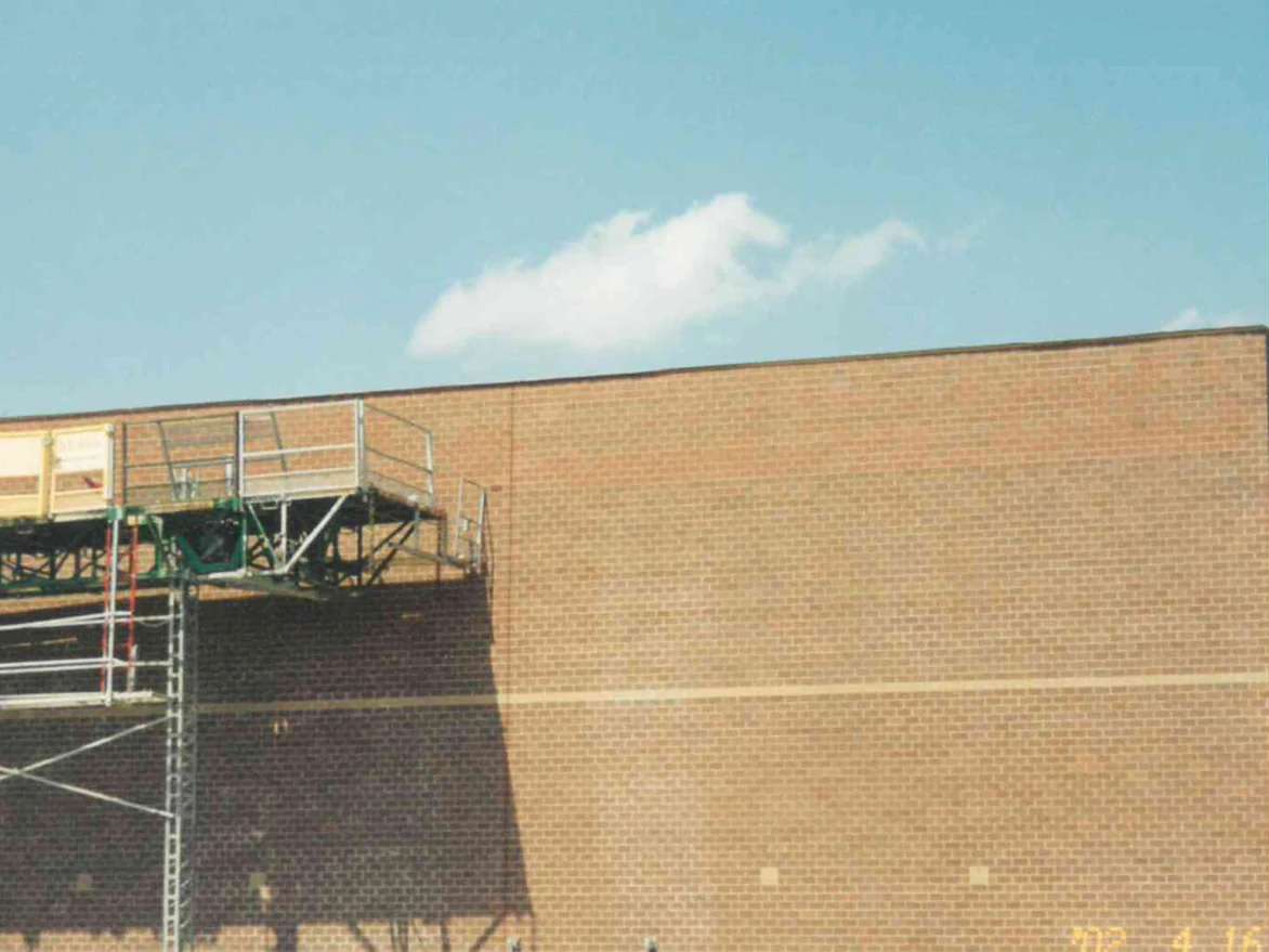 A work platform on a mast climber against the school wall in April 2002, with a lighter run of brick along the top of the elevation above a single buff accent course
