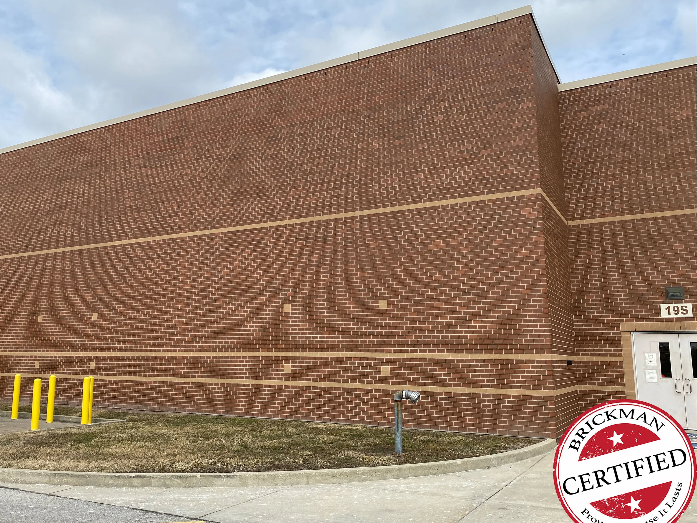 The side wall of the school in 2021 from the parking lot, red brown brick at one range from the ground to the parapet, two buff accent courses, yellow bollards and a door marked 19S at the far end, with the certification stamp in the corner