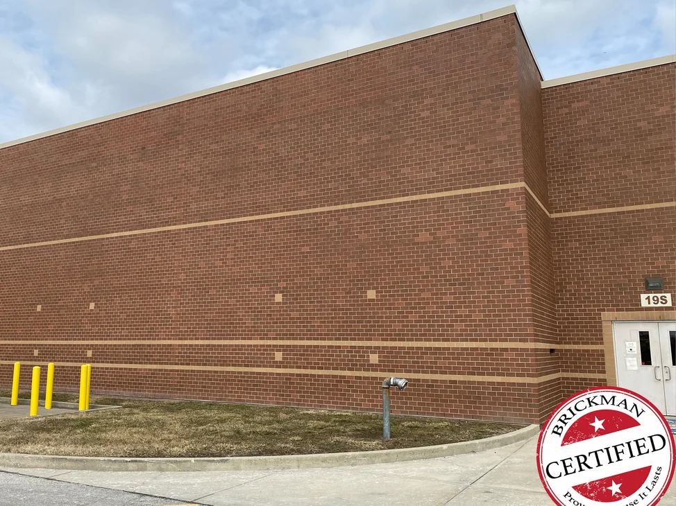 The side wall of the Indianapolis school in 2021, red brown brick at one range with two buff accent courses, yellow bollards at the near end and the certification stamp in the corner.