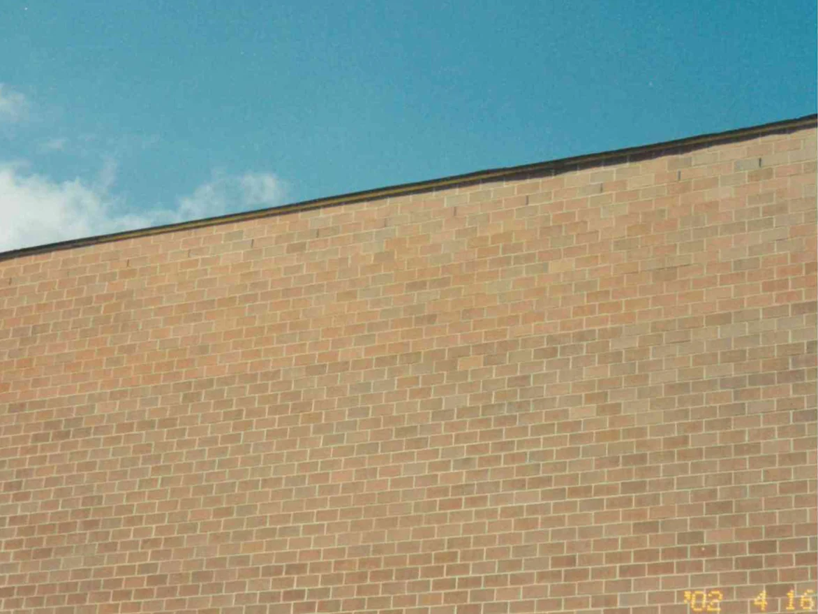 The upper part of the school wall in April 2002 under a dark parapet cap, a run of pale tan brick across the top with the brick below it reading darker and browner