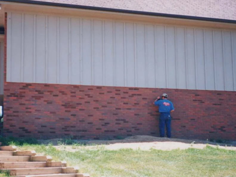 An applicator in a blue shirt working stain into the red brick base of the addition's side wall under tan board-and-batten siding, a second man seated on the bank at left