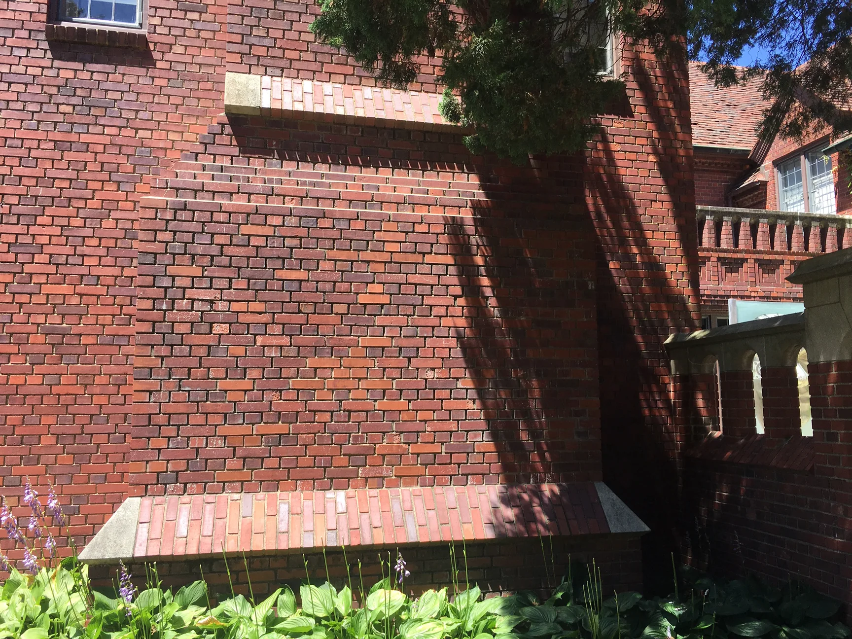 Close view of the chimney wall in 2019, reds and browns running unit to unit under a stone-capped offset with hostas at the foot