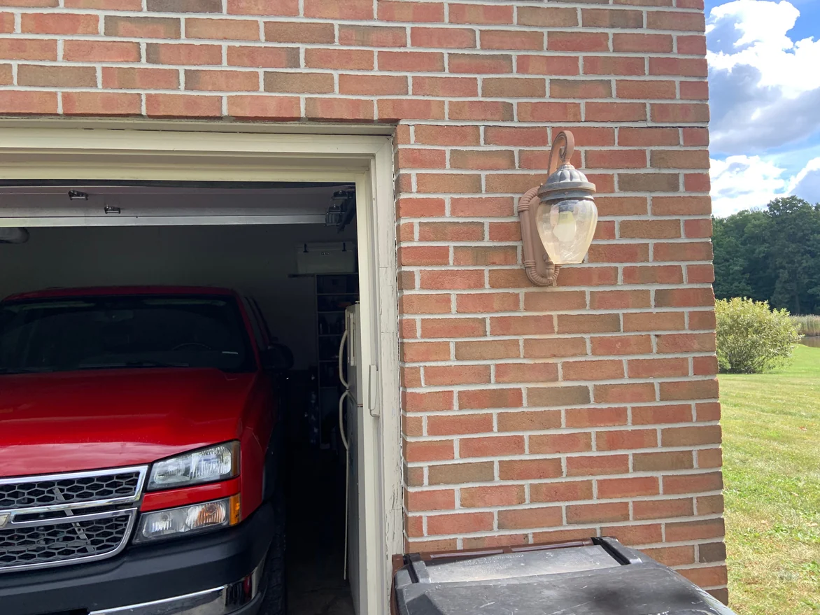 The outside corner of the attached garage in 2019, a carriage lamp on blended brick with the lawn and treeline beyond