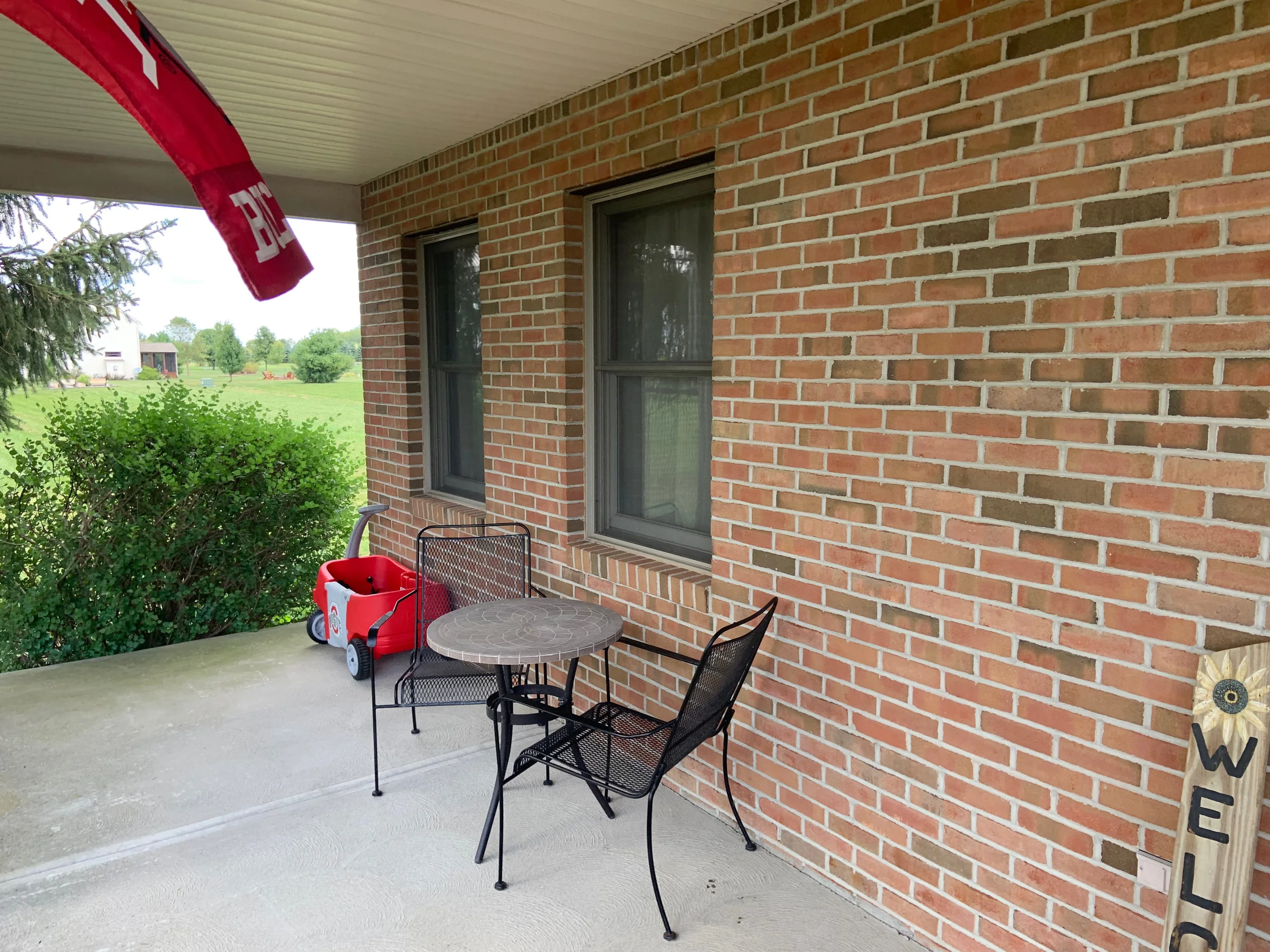The covered porch in 2019 looking out to the lawn, a table and chairs against a brick wall that reads as one even red and tan range past two sash windows