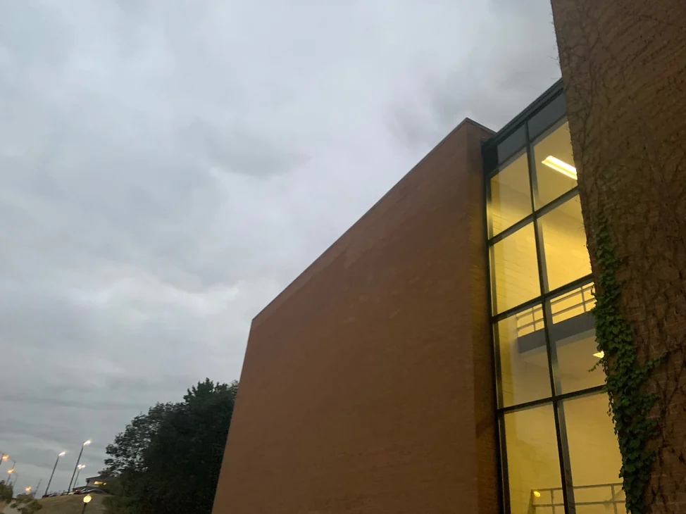 The end wall of the university building in 2019, seen from an angle at dusk, brown brick running to a lit glass stair bay.