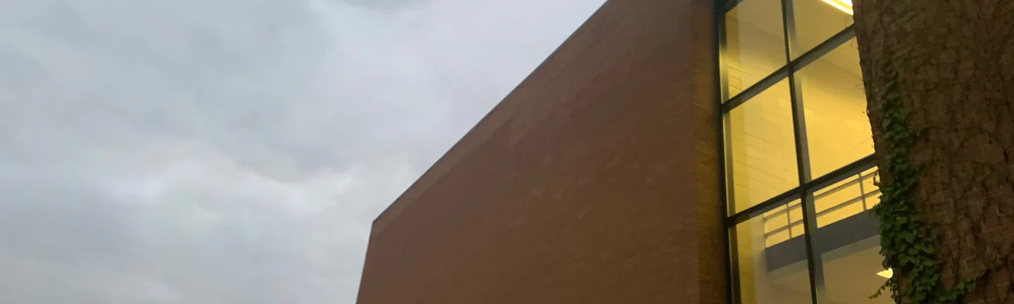The end wall of the university building in 2019, seen from an angle at dusk, brown brick running to a lit glass stair bay