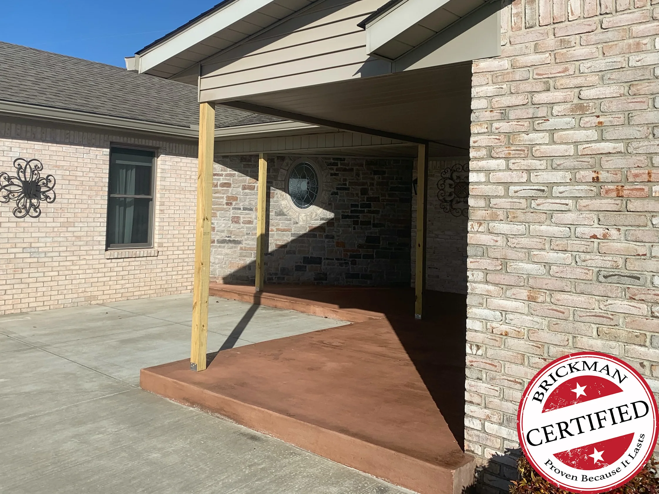 The covered patio in 2020 under a clear sky, the pale brick wall on the left and the whitewashed-look brick of the gable wing on the right meeting at new timber porch posts over a stained concrete step, with the certification stamp in the corner