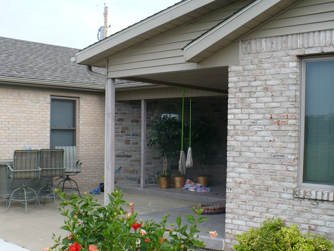 The covered patio in 2008 before staining, the wall on the left a uniform cream brick and the gable wing on the right a whitewashed-look brick with pink flashing, patio chairs and a hibiscus in front