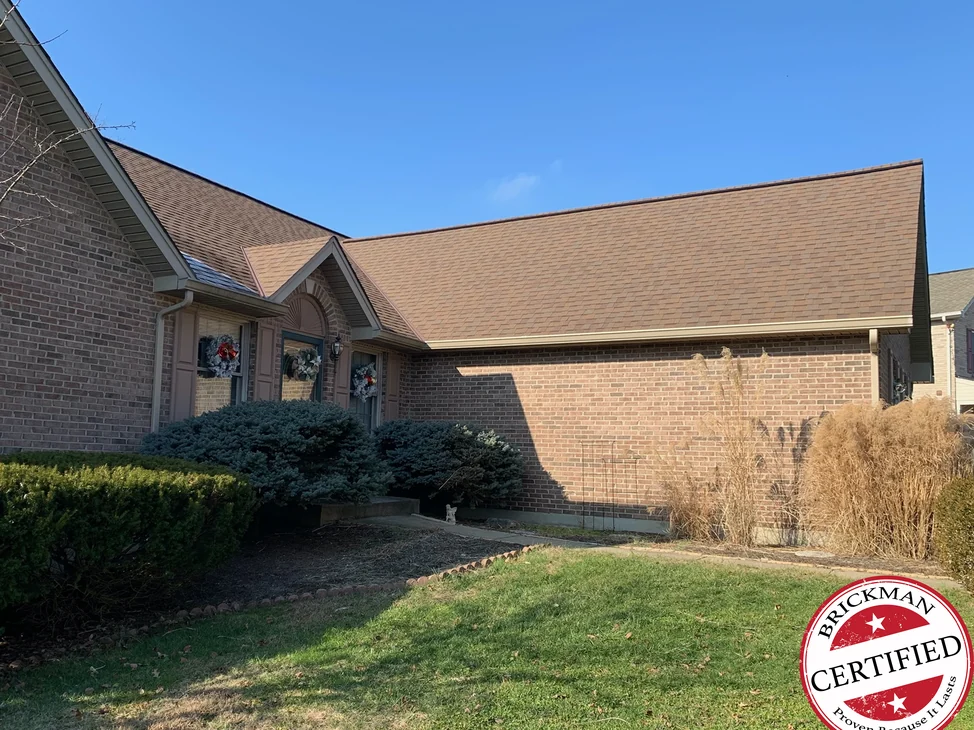 The front of the house in 2020 under a clear sky, the entrance gable and the long wing beyond it in one even red brown brick, with the certification stamp in the corner.
