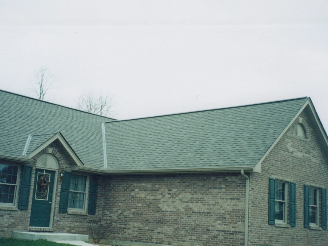 The front of the house in March 1999 under an overcast sky, pale blotches of light brick scattered through the grey brown wall between the entrance gable and the two shuttered windows