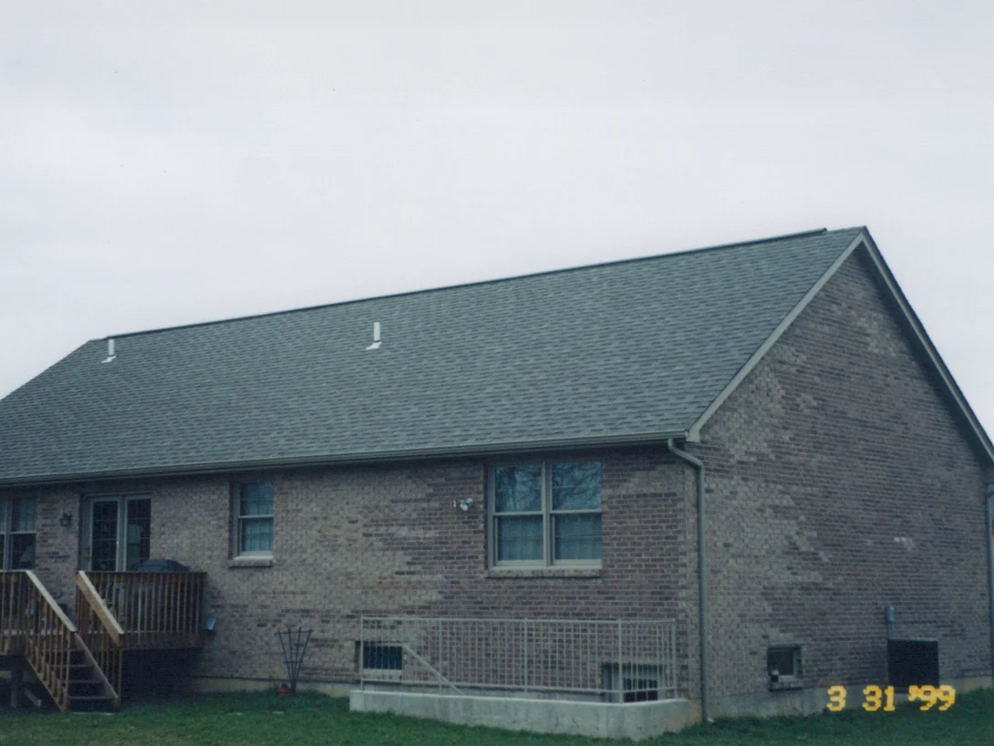 The rear of the house in March 1999, a wood deck at the left and a walk-out basement wall below, with patches of light brick across the long wall and the gable end