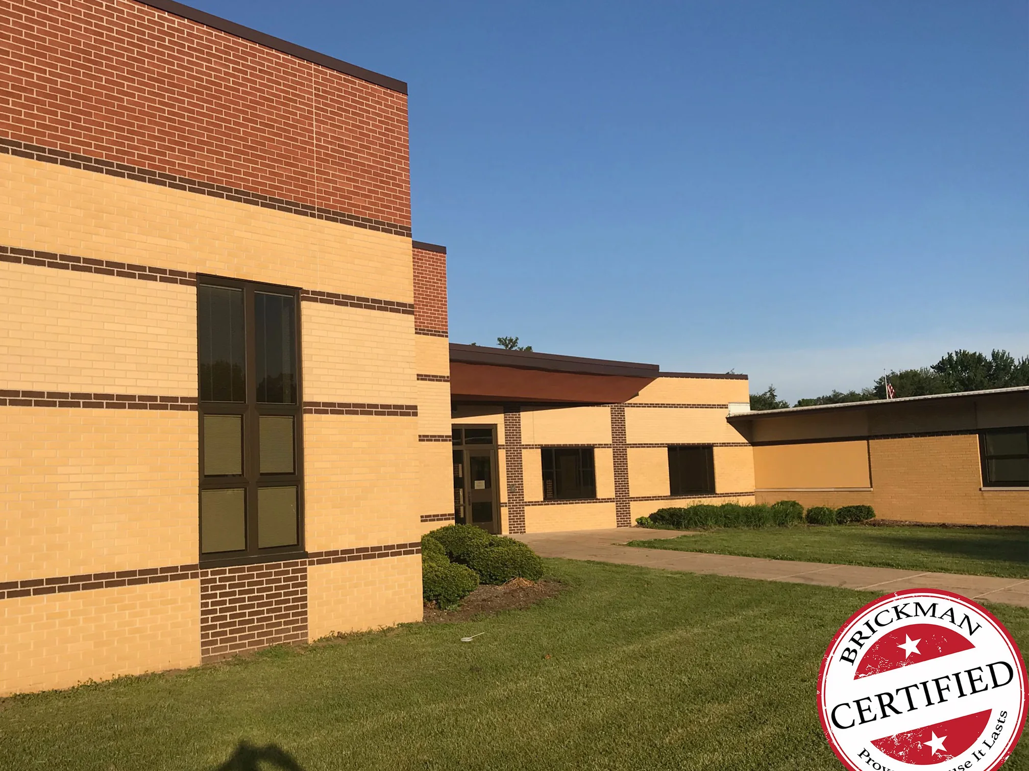 The taller wing and the older single-story range meeting across a lawn in 2019, buff brick below and a band of red brick above, with the Brickman certified stamp in the corner of the frame