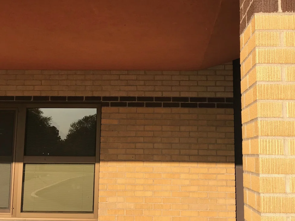 Close view under the entrance canopy in 2019, individual buff brick units above a window with a dark brown course running between them and a brick pier at the corner
