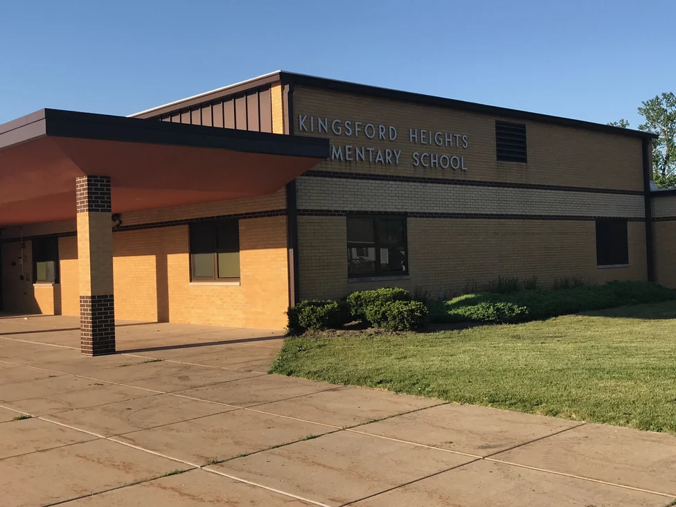 The entrance end of the school in 2019 read at an angle, KINGSFORD HEIGHTS ELEMENTARY SCHOOL in metal letters on the buff brick above a dark brown banding course