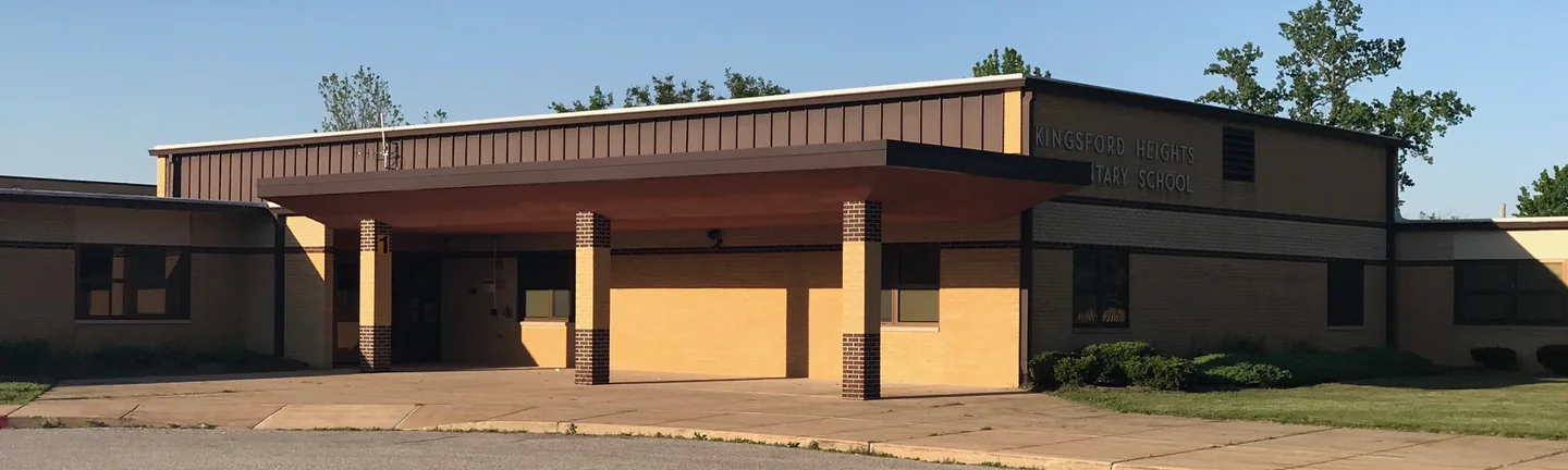 The front of a low school building in 2019, its buff brick running unbroken past the entrance canopy