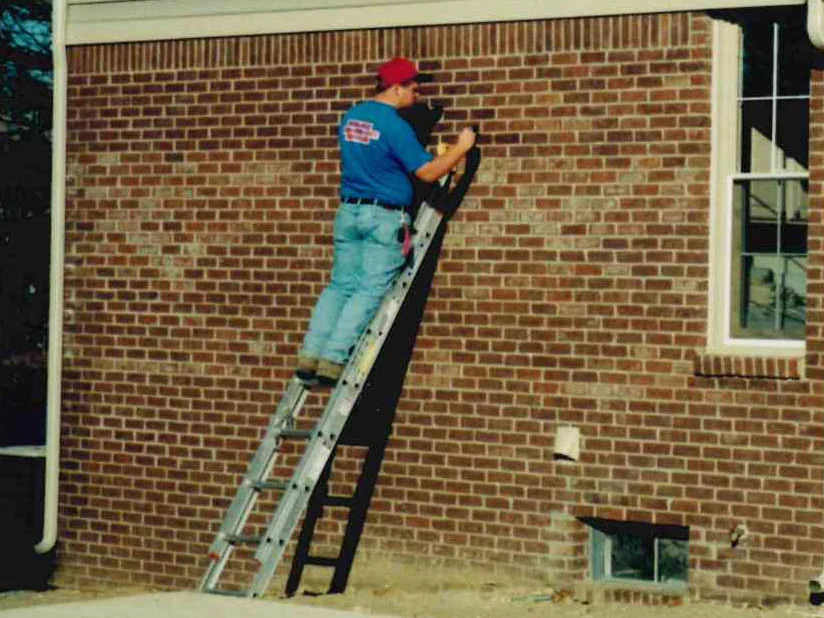 The applicator on the ladder in 1999 seen from the drive, staining brick beside a basement window well, the wall a mix of dark and pale units