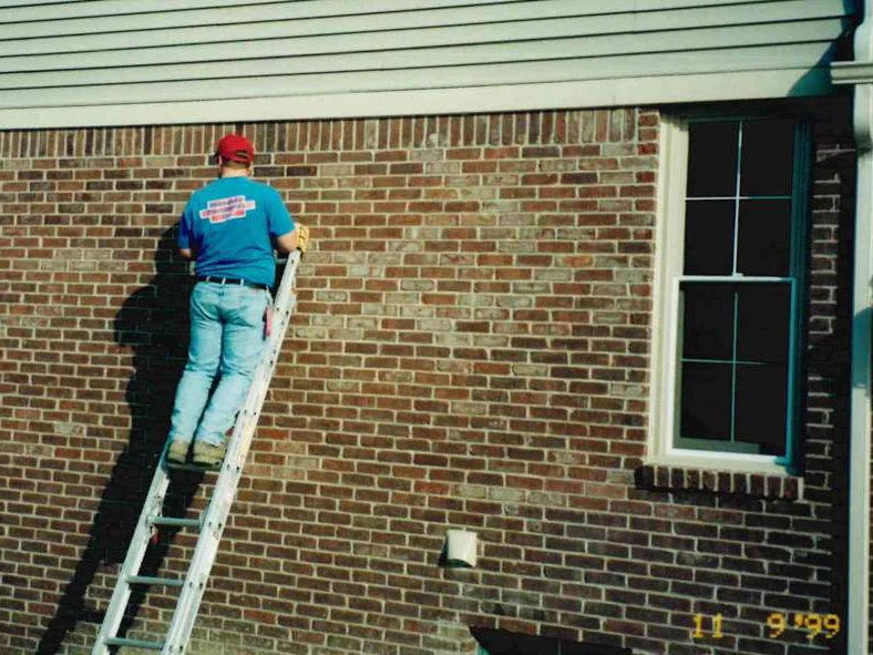 An applicator on an extension ladder in 1999, working stain into the brick of a side wall below a band of siding, a window to the right and a camera date in the corner