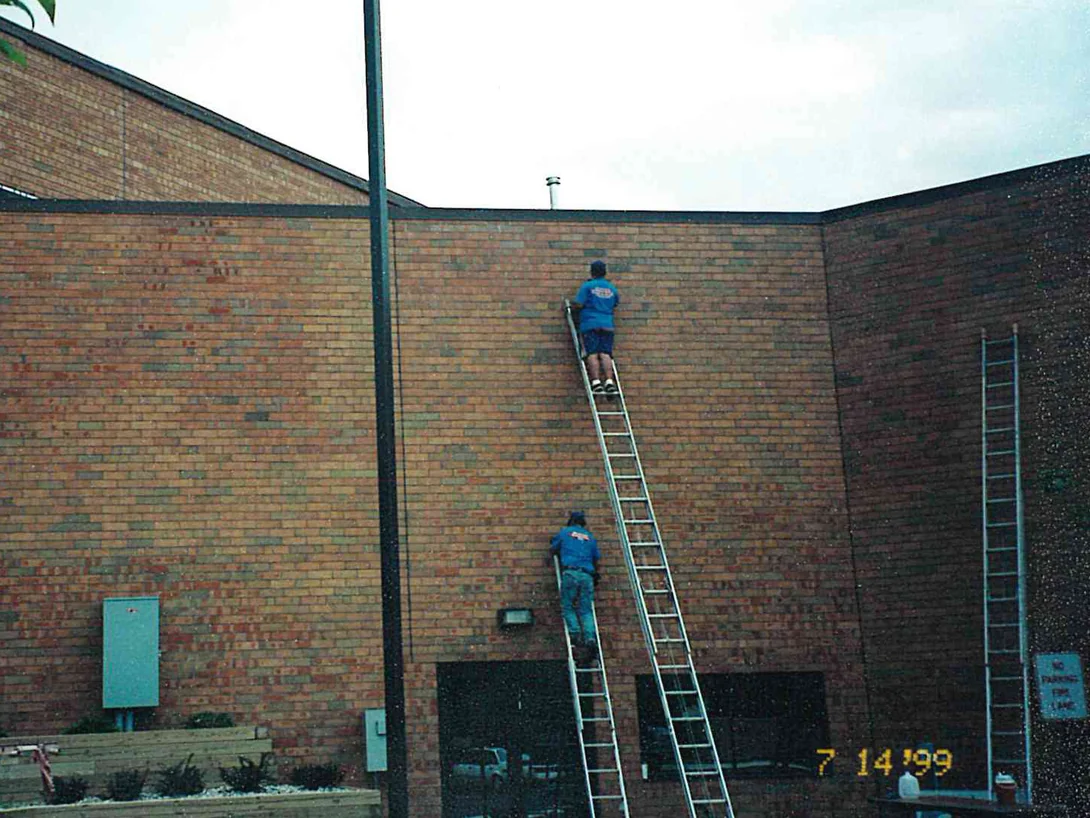 Two applicators on ladders working across a tall blank brick wall at an inside corner of the office in 1999