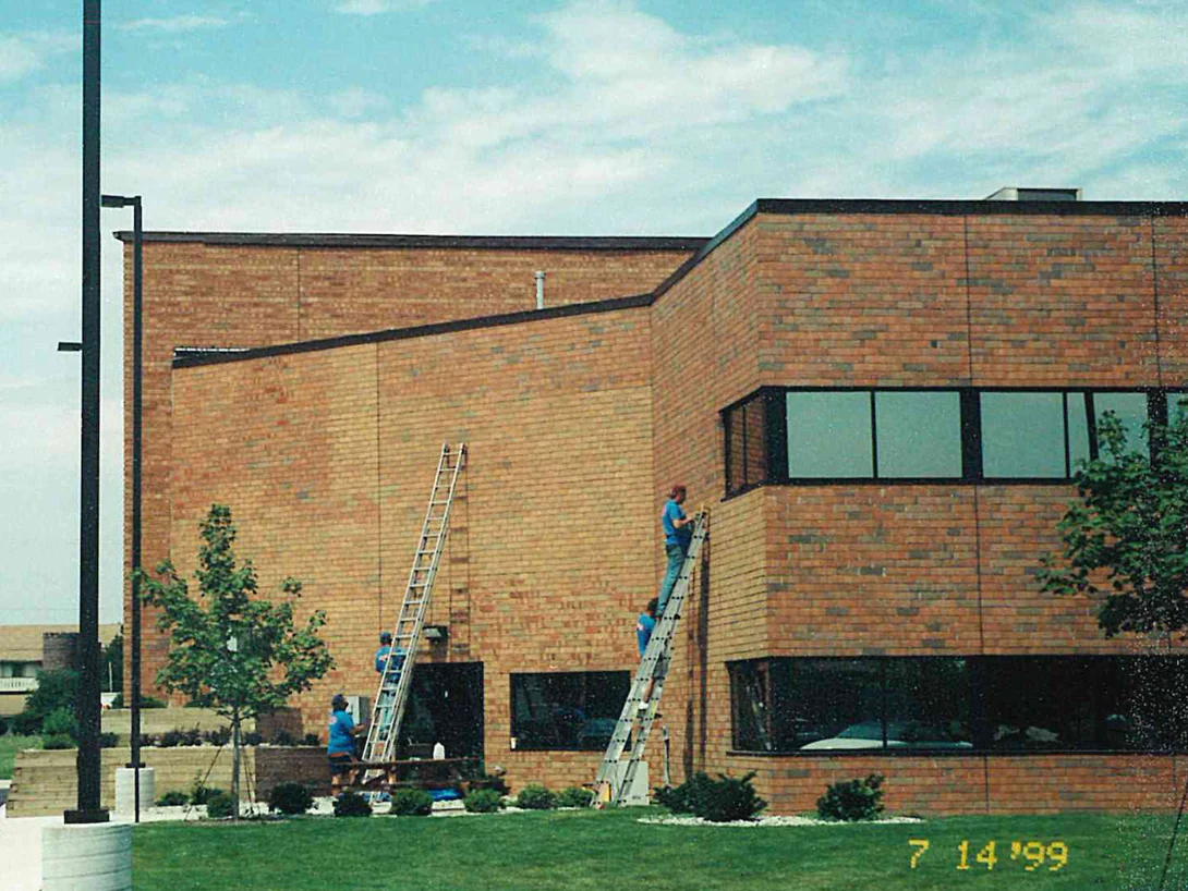 A two-story office building in 1999 with applicators on ladders at its corner, the brick a pale and uneven orange and tan