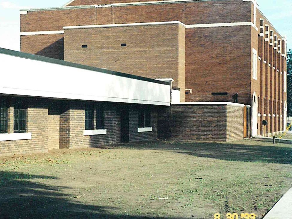 A new low school wing in pale tan brick standing in front of a taller block in darker brown brick, on bare graded ground