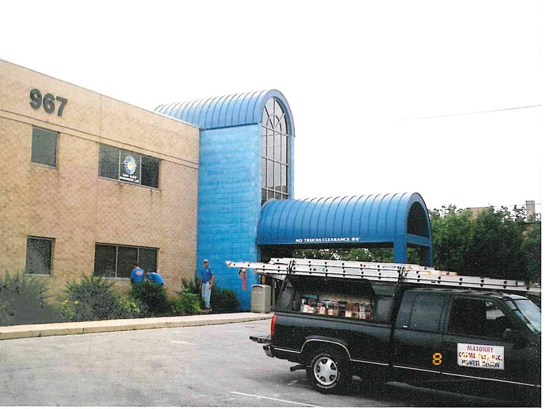 The two-story buff brick office building in 2003, its blue tile entrance tower and barrel awning beside a crew pickup with ladders on the rack