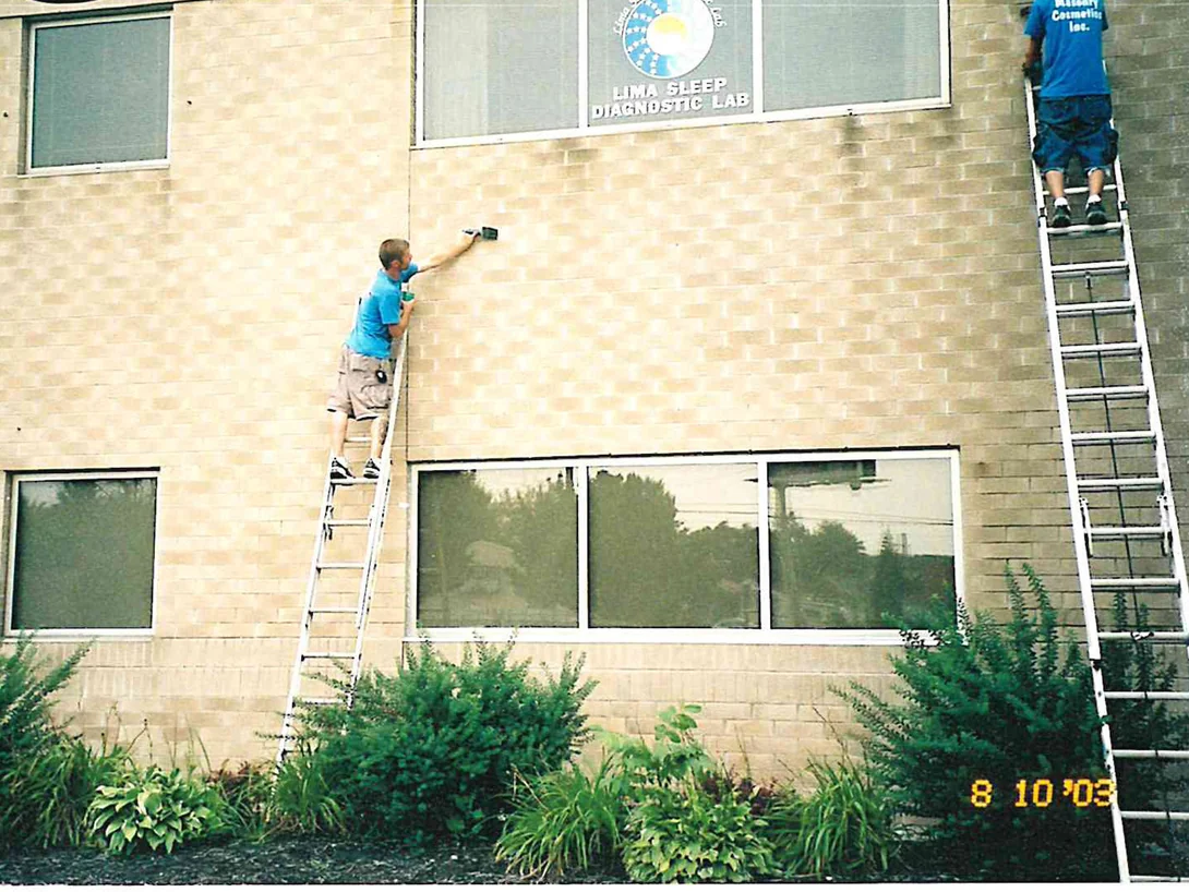 Two applicators on extension ladders in August 2003, one working a hand brush over single buff brick units below the tenant sign, with a burned-in camera date in the corner
