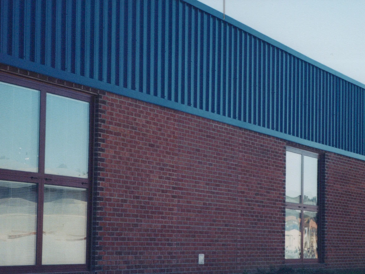 The long red brick elevation of the high school in 2000, three window bays under dark blue corrugated metal cladding, with a small utility box low on the wall between the first two windows