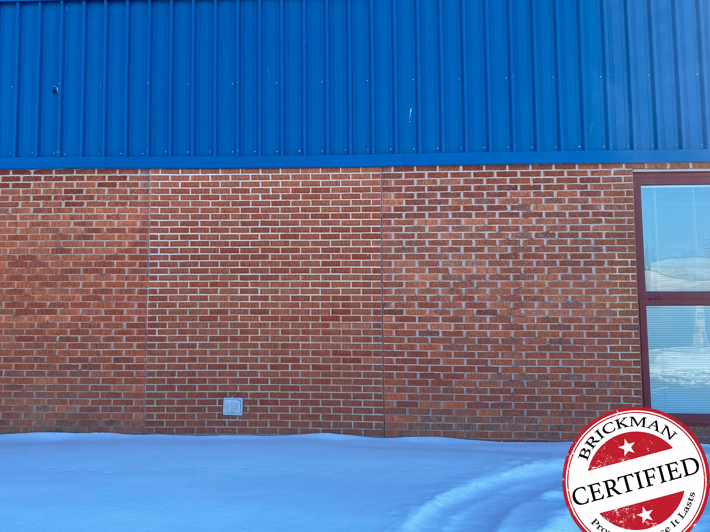 The same stretch of wall in 2021 in snow, the filled-in panel between its two joints reading as one red range with the brick beside it under bright blue metal cladding, the utility box still at its foot and the certification stamp in the corner