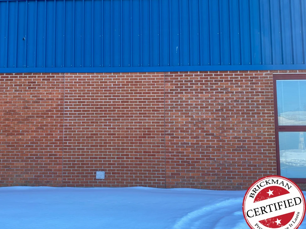 The red brick wall of the high school in 2021 in snow, the filled-in panel between its two joints reading as one range with the brick beside it under bright blue metal cladding, with the certification stamp in the corner.