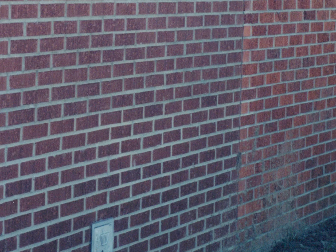A close view of the filled-in panel in 2000, its darker brown brick set between two vertical joints with the brighter red original brick on either side and a utility box at its foot, an extension ladder leaning in at the right