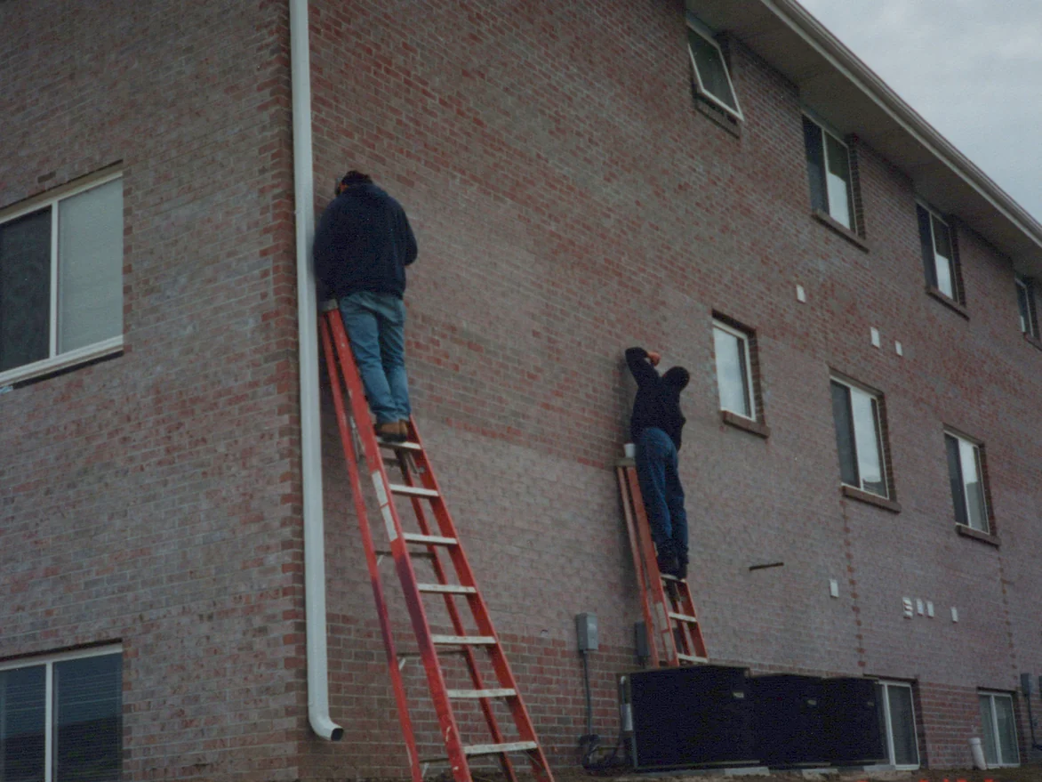 Two applicators on extension ladders working stain into the brick at a corner of the building in 1997, a downspout between them and three rows of windows running off to the right