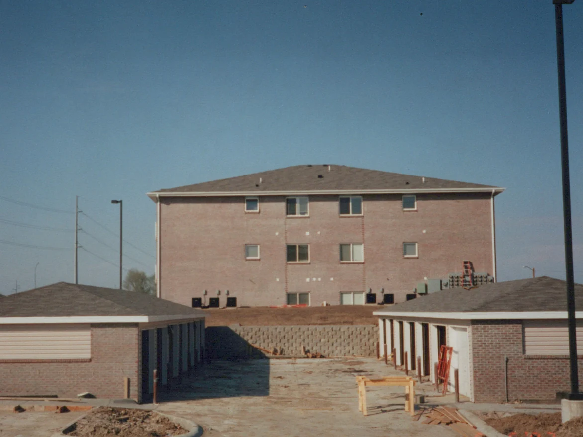 The three-story apartment building in 1997 seen across an unfinished garage court, pink brick under a hipped roof with a retaining wall and bare dirt in front, a camera date in the corner