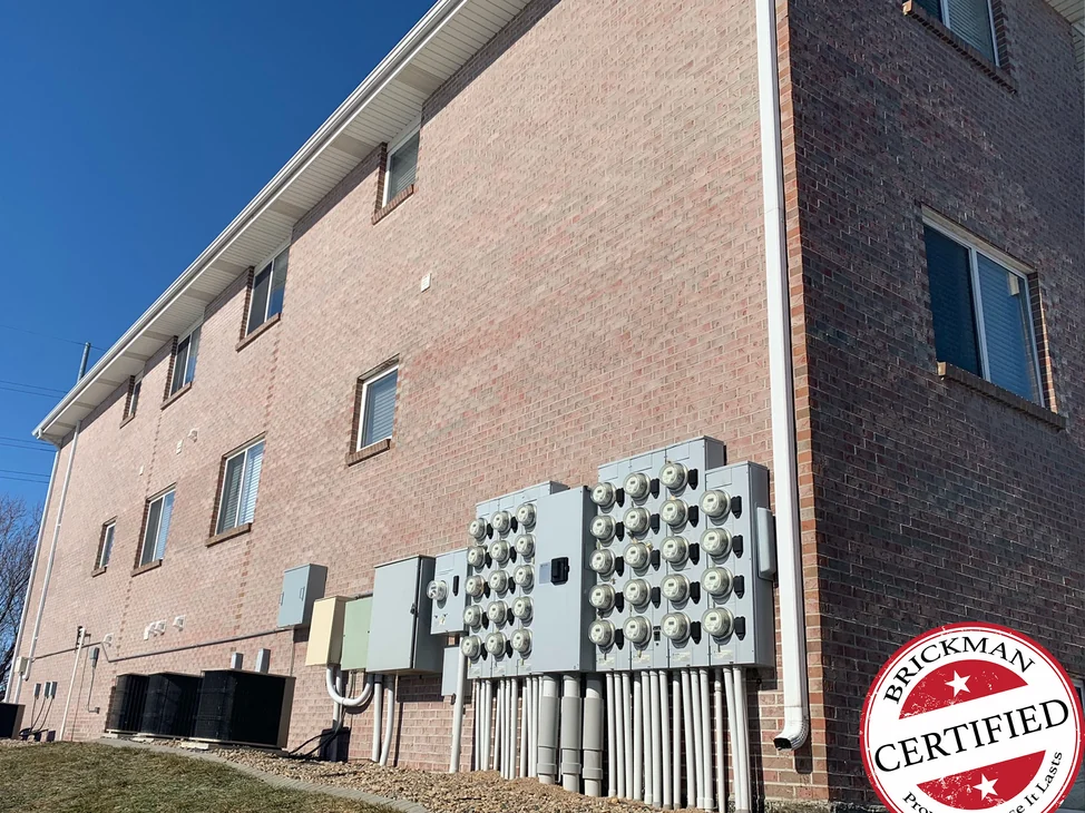 The long side wall of the apartment building in 2021, pink brown brick in direct winter sun above a bank of electric meters, with the certification stamp in the corner.