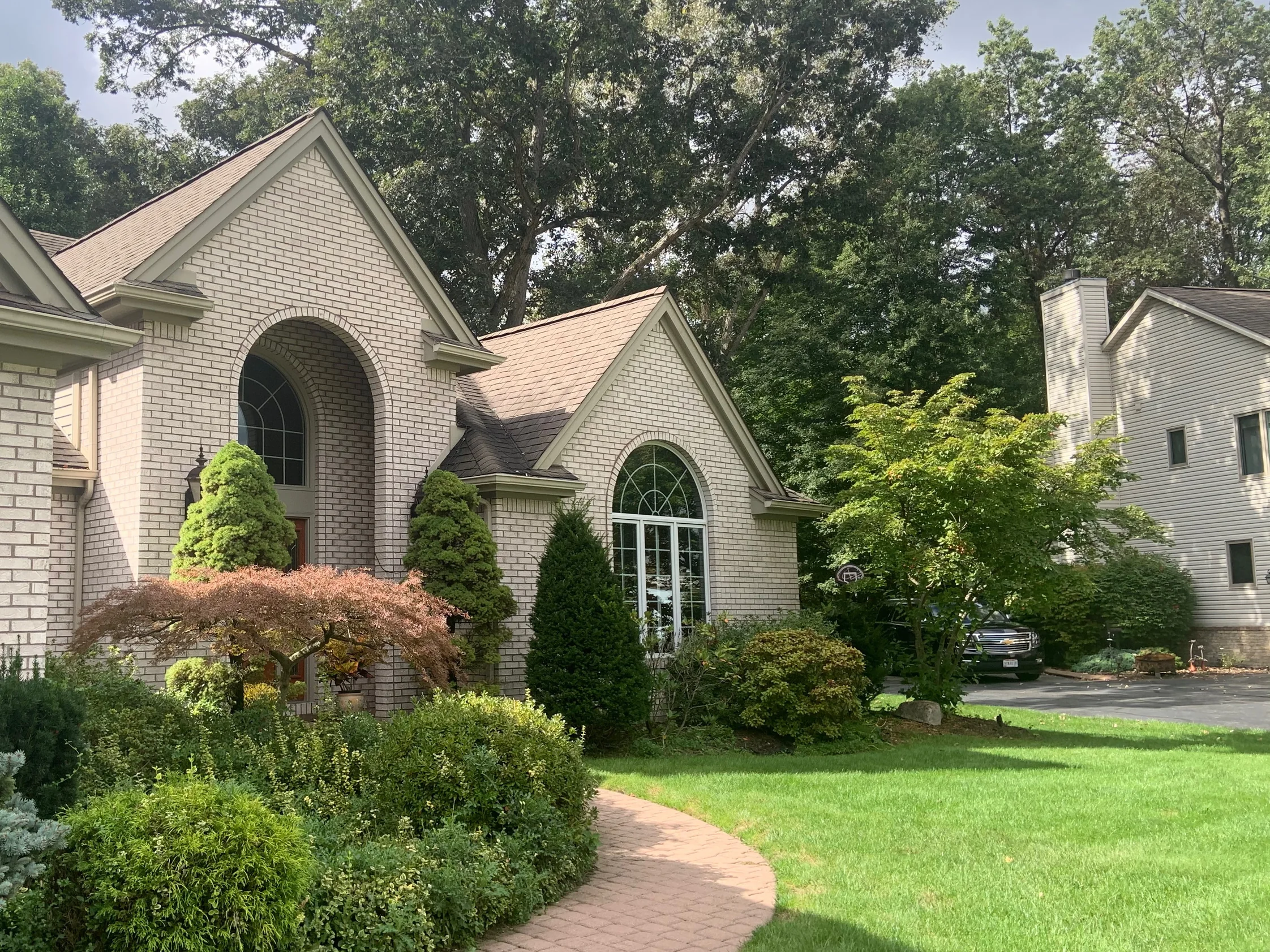The front of the house in 2019, an arched entry gable and a second gable with a fanlight window in one pale cream brick, mature planting along a paver walk and lawn