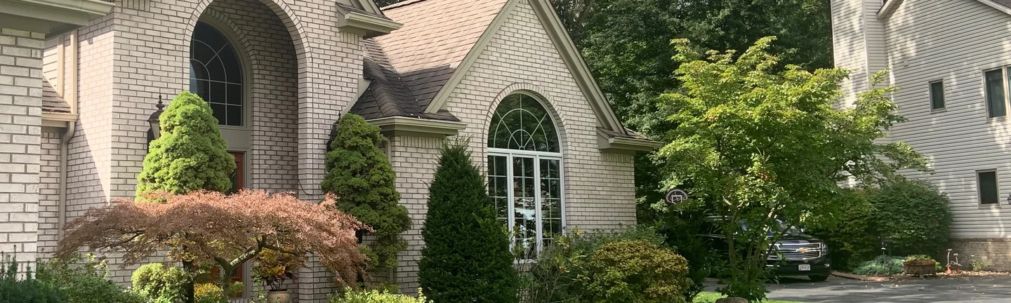 The front of the Lost Creek house in 2019, an arched entry gable of pale cream brick behind planting, with a second gable and a neighboring house beyond