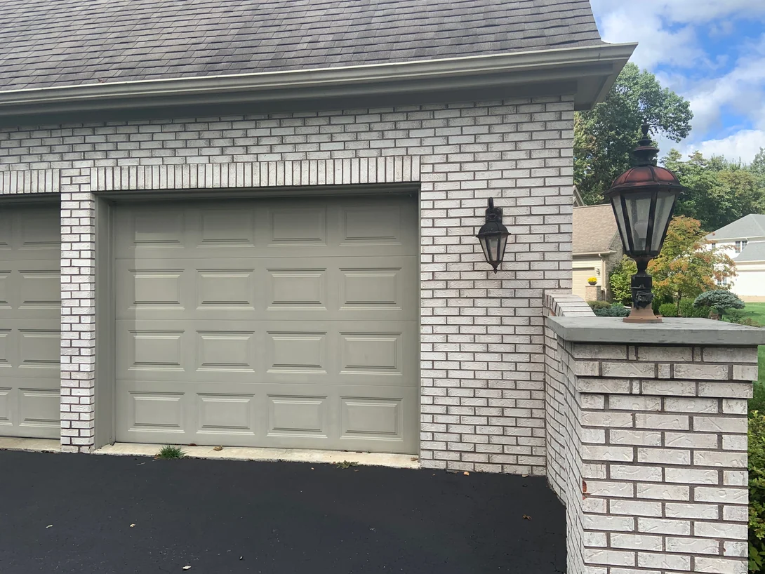A garage door and a brick pier with a lamp post in 2019, the cream brick and its soldier course above the door one color with the house behind
