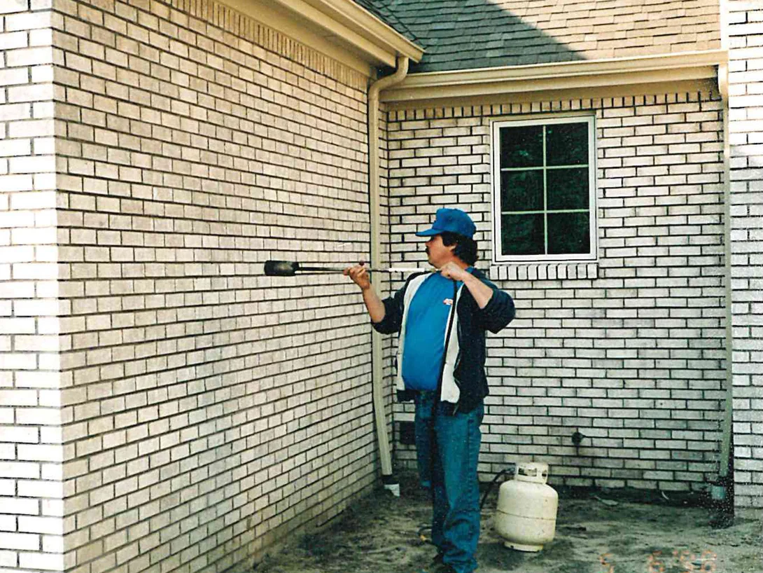 An applicator in a blue cap working stain into the cream brick wall with a brush on a long handle in 1998, a propane cylinder on the ground beside him