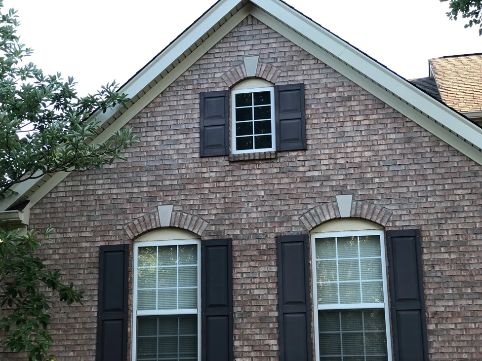 The upper gable in 2019 at close range, an arched window with shutters set in blended brick under a soffit and a brick arch head